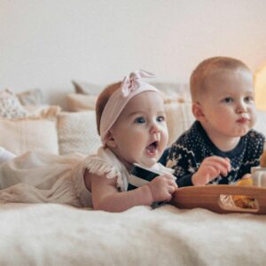 Adorable siblings enjoying a cozy time at home, surrounded by cushions and snacks on bed.
